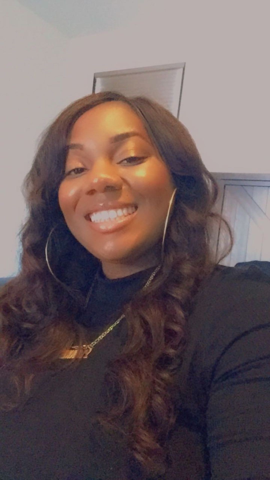 Woman smiling while taking a selfie indoors, wearing a black top and hoop earrings.